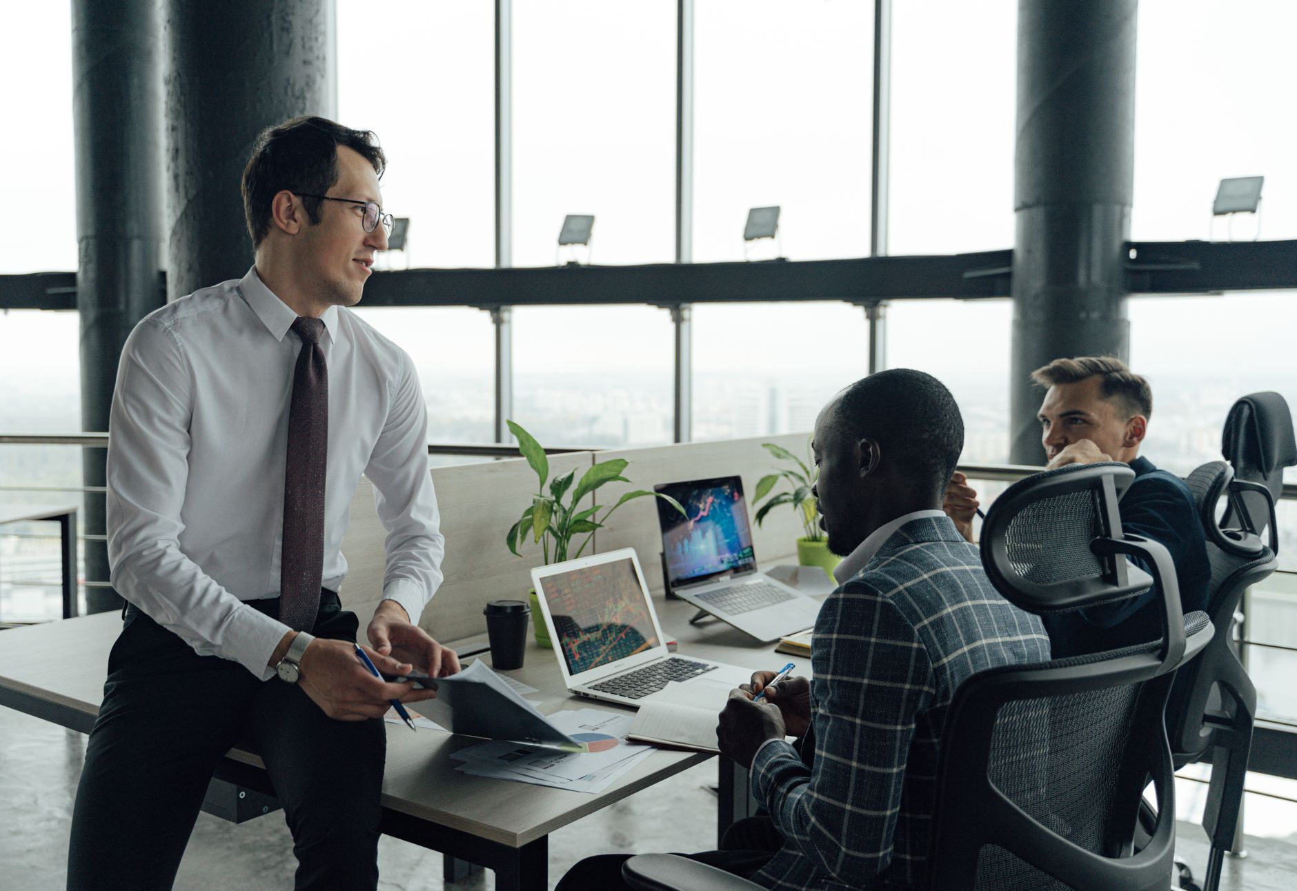 Three men engaged in a business meeting in a modern office setting with laptops and documents. — How Can One Save Money While
