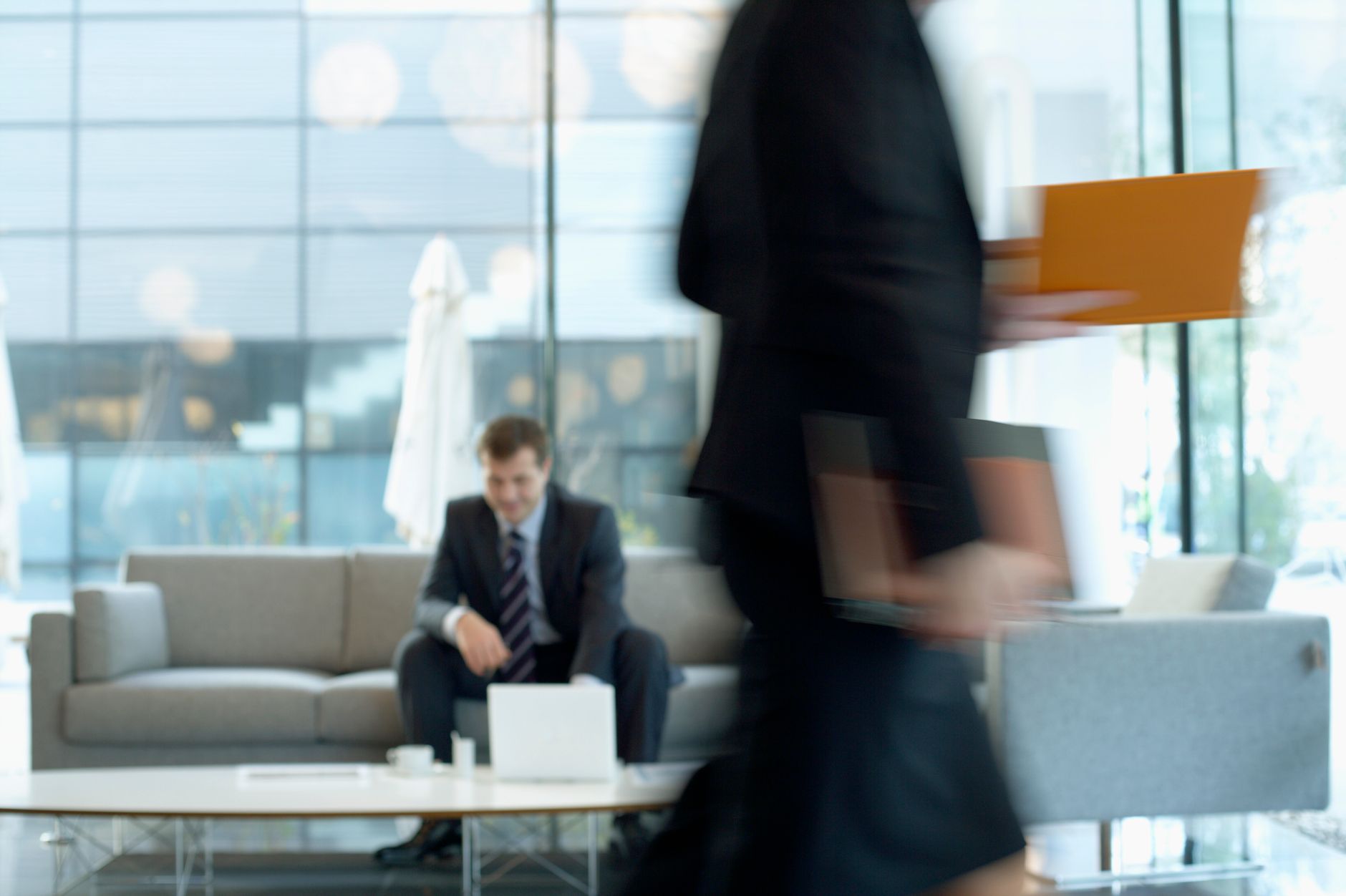 Businessman sitting in modern office lounge using a laptop while colleague walks past, creating dynamic blur. — International